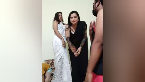 Video of a woman in a white lace wedding gown standing beside a woman in a black lace dress, with a man's arm visible in the right foreground.