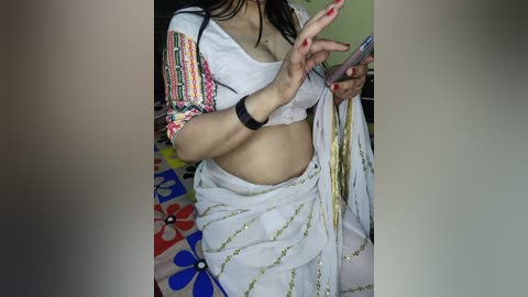 Video of a South Asian woman in a white sari with colorful embroidery, holding a phone, against a floral-patterned bedspread.