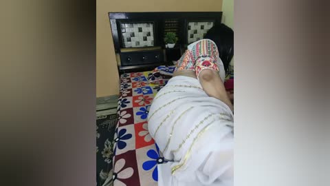 Video of a person lying on a bed with a colorful floral bedspread, wearing a white robe with gold trim and multicolored slippers. The room has a dark wooden headboard with a mosaic design, and a black and white patterned carpet.