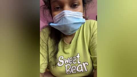 Video of a South Asian woman with medium brown skin, wearing a light yellow t-shirt with \"Sweet Bear\" in pink text, a blue surgical mask, and a pink headband. She has curly black hair and is lying on a pink bed.