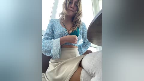 A video of a young woman with blonde hair, wearing a teal bra and a light blue floral top, sitting on a white chair with a beige skirt, in a bright, modern room.