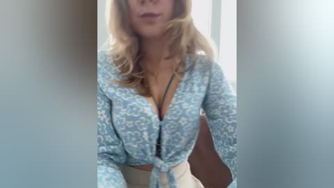 A video of a blonde woman with light skin, wearing a blue floral-patterned robe tied at the waist, partially revealing a black bra. The background is a blurred, indoor setting with neutral colors.