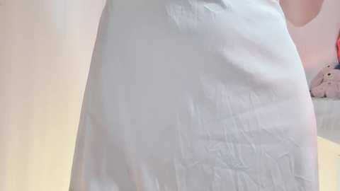 Video of a person in a white hospital gown, with slight creases, standing in a room with light pink walls and a plush toy in the background.