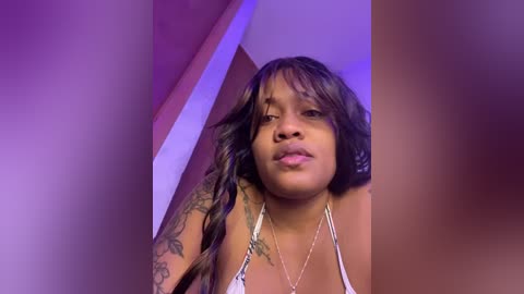 Video of a Black woman with long, wavy hair, wearing a white, floral-patterned bra, standing indoors under purple lighting. Her face is partially blurred.