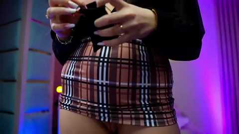 Video of a woman in a tight, short plaid skirt, holding a camera, in a dimly lit room with purple and blue lighting.