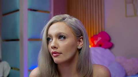 Video of a young woman with straight, platinum blonde hair and light skin, wearing a strapless top, gazing thoughtfully. Background features a colorful room with soft lighting, including a pink flower and a bed with a fluffy white blanket.