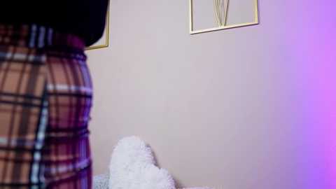 A video of a cozy, minimalist room featuring a beige wall with a gold-framed abstract painting, a plush white throw, and a plaid blanket on a bed.