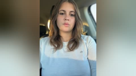 Video of a young woman with light skin and shoulder-length brown hair, wearing a white and blue striped long-sleeved top. She's inside a car, with blurred background.