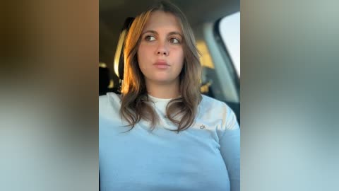 Video of a young woman with long, wavy brown hair, wearing a white long-sleeve shirt, looking contemplative with a slight frown, sitting in the backseat of a car.