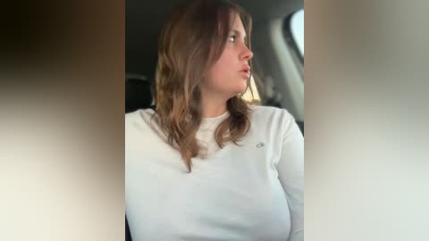 Video of a young woman with wavy, shoulder-length brown hair, wearing a white long-sleeve shirt, seated in a car. She gazes out the window, looking thoughtful. Background includes car interior details and sunlight.