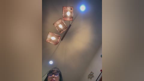 Video of a dimly lit room with a modern, triangular chandelier featuring three amber-colored, square lanterns with glowing white bulbs. The ceiling is beige, and a person with glasses and dark hair is partially visible at the bottom.
