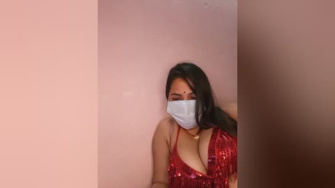 A video of a young woman with long black hair, wearing a red sequined dress, a white face mask, and a gold necklace, seated against a plain, light-colored wall.