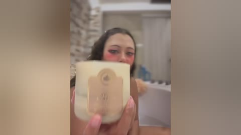 Video of a young woman with fair skin, dark hair, and a slight blush, holding a beige candle in a bathroom with beige walls, white sink, and a partially visible shower.