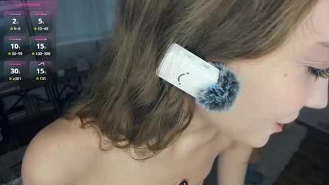 Video of a young Caucasian woman with shoulder-length brown hair, wearing a white earbud with a blue furry earmuff, in a dimly lit room.