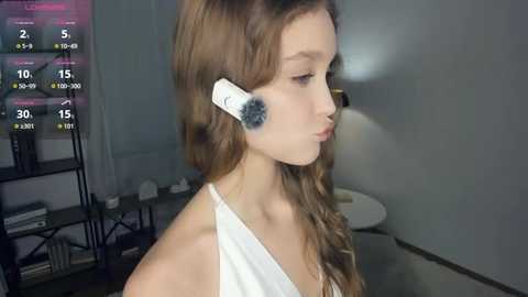 A video of a young woman with long, wavy brown hair, wearing a white off-shoulder top, with a white headphone with a black microphone attached to her ear, standing in a dimly lit room with a gray wall and a table in the background.