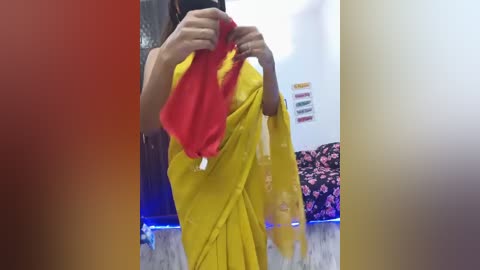 Media: Video of a person wearing a yellow sari with red borders, holding a red scarf, in a colorful room with a flowered bedspread and wall art.