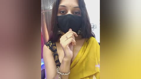 Video of a young South Asian woman with medium skin tone, wearing a yellow saree, black mask, and black floral-patterned blouse. She is indoors with a blurred background.