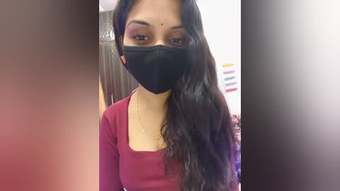 Video of a young woman with long, wavy black hair, wearing a black face mask, maroon top, and gold necklace, in a bathroom with a tiled wall and colorful nail polish bottles in the background.