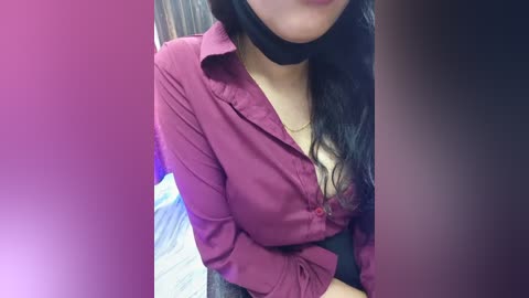 A video of a woman with long black hair, wearing a partially unbuttoned maroon blouse, revealing a black lace bra, seated indoors with blurred background.