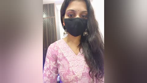 Video of a South Asian woman with long black hair, wearing a pink traditional sari with intricate white embroidery, black mask, and large silver earrings, standing indoors with blurred background.