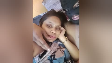 A candid video of a woman with medium brown skin, short black hair, and a floral blouse, resting on a bed with a striped pillow.