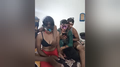 Video of three people in a room with white walls; a woman in a black bra and red skirt, a man in a green shirt, and another man in a mask, holding a phone.