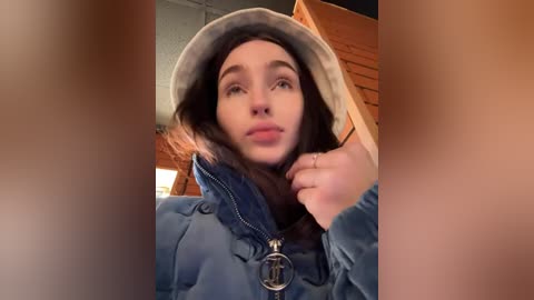 Video of a young woman with fair skin, wearing a gray beanie, blue jacket, and necklace, looking thoughtful in a brick-walled room with warm lighting.