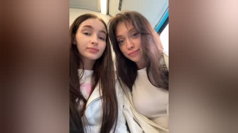 Video of two young women with long, straight brown hair, wearing white shirts, smiling, seated on a plane.