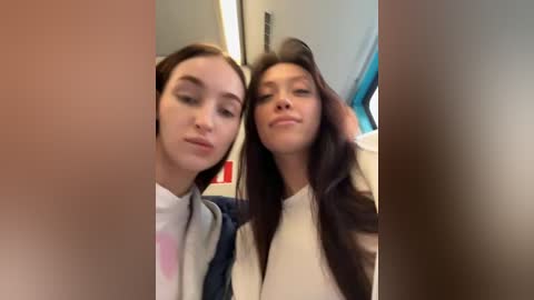 Video of two young women with fair skin and long dark hair, smiling and leaning close together, seated on a train. Background shows train interior with blue seats and gray walls.