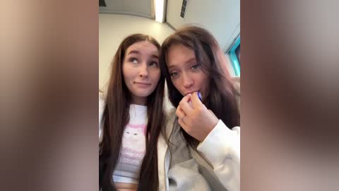 Video of two young women, one with long brown hair, wearing a white crop top, and the other with long brown hair, wearing a light jacket, holding a purple lollipop. They are smiling inside a car.
