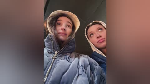 Video of two young women with light skin, wearing hooded jackets, looking up at something out of frame, in a dimly lit room.