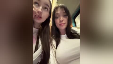 Video of two young women with fair skin and long brown hair, wearing white tops, posing closely in an indoor setting with blurred background.