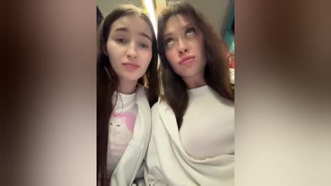 Video of two young Asian women with long, dark hair, wearing white tops, posing closely together, slightly smiling, in a dimly lit room.