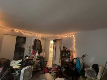 A video of a messy, cluttered bedroom with a white door open, exposing a chaotic interior. Walls are adorned with fairy lights, and a bicycle is partially visible.
