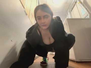A video of a woman in a black top and leggings, squatting in a narrow, dimly-lit hallway with white walls and wooden floor. She has medium skin tone and long brown hair.