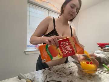 A video of a curvy, fair-skinned woman with brown hair in a kitchen, pouring orange juice from a carton onto a marble countertop.