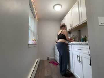 Video of a woman in a small, narrow kitchen with light grey walls and white cabinets. She is wearing a black bra and dark pants, cleaning the countertop. A pink towel lies on the floor.
