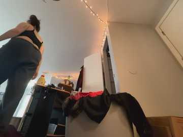 Video of a woman in a black sports bra and pants, leaning over a white refrigerator, surrounded by kitchen clutter and string lights.