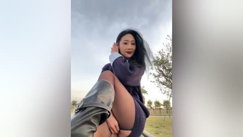 Video of an Asian woman with long black hair, wearing a purple sweater dress, black boots, and sitting outdoors against a cloudy sky with trees in the background.