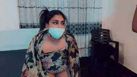 Video of a woman with medium skin tone, wearing a floral-patterned blouse and light blue surgical mask, seated indoors with beige curtains and dark furniture in the background.