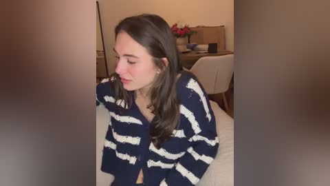Video of a young woman with long dark hair, wearing a blue and white striped cardigan, sitting on a bed in a dimly lit bedroom with a desk, chair, and flowers in the background.