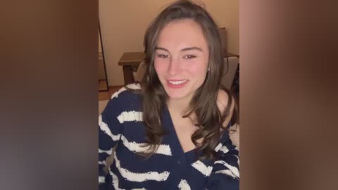 Video of a smiling, light-skinned woman with long, wavy brown hair, wearing a navy blue and white striped sweater. Background includes a wooden table and beige walls.