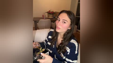 Video of a young woman with long, wavy dark hair, wearing a navy blue and white striped sweater, sitting in a modern living room with beige furniture, holding a glass of wine.