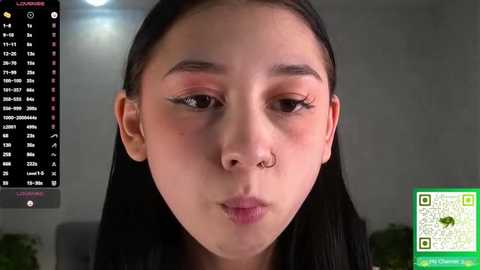 Video of an Asian woman with long black hair, light skin, and a small nose ring, making a kissing gesture. Background includes a calendar and green QR code.