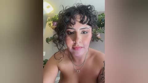 Video of a young woman with curly black hair, light skin, and a nose piercing, wearing a heart necklace, standing topless in a room with green plants and a ceiling light.