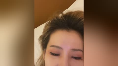 Video of a woman with fair skin and dark brown hair, resting on a white pillow. Her eyes are closed, and she wears subtle makeup with dark eyeliner.