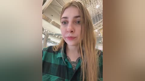 A video of a young woman with long, straight blonde hair, wearing a green and black plaid shirt, standing indoors near a chandelier.