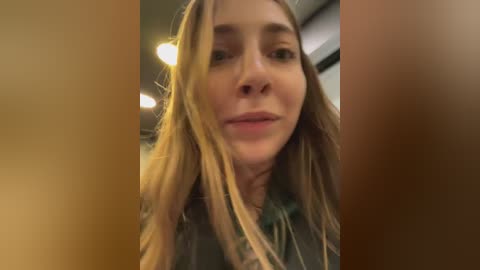 A close-up video of a young woman with long, straight, blonde hair and light skin, wearing a dark jacket. The background is blurry, suggesting an indoor setting with warm lighting.
