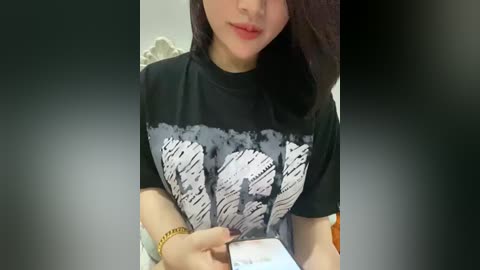 Video of an Asian woman with fair skin, long dark hair, wearing a black graphic t-shirt with white \"POTUS\" text, holding a smartphone. Background is blurred, showing a white wall.