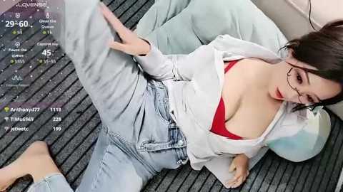 A video of a young Asian woman with glasses and fair skin, wearing a red bra and light-wash denim jeans, reclining on a gray-striped carpet.